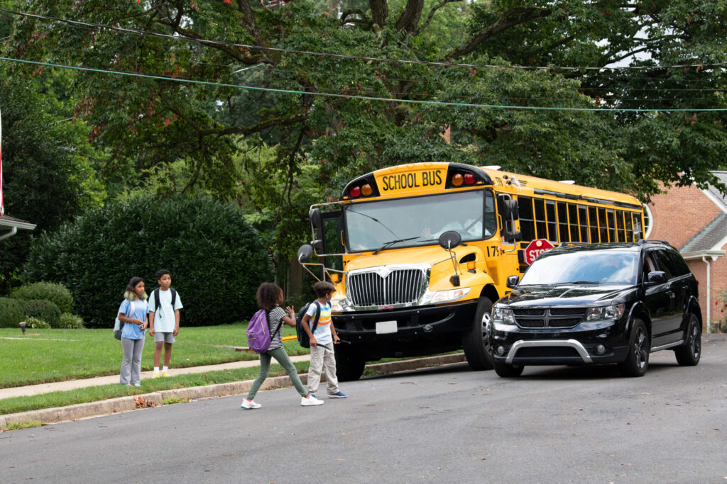 Alt text: “Children cross near a stopped school bus as a passenger vehicle occupies the roadway in a staged safety scene.”