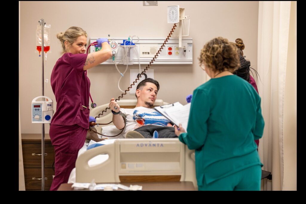 A healthcare worker adjusts equipment while another interacts with a patient in a hospital bed.