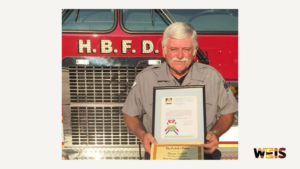 Danny Coffelt holding a framed certificate in front of a Hokes Bluff fire truck, with the WEIS logo.