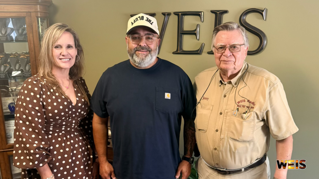 Loblolly Fest announcement photo showing three people posing at WEIS Radio with the station’s wall sign behind them.